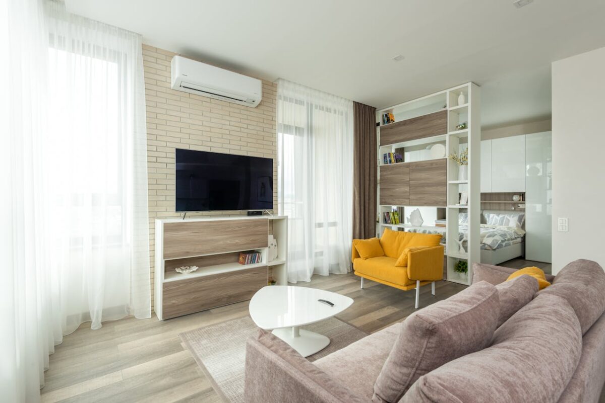 who-we-are Cozy sofa and armchair placed near cabinet with TV set in contemporary living room separated from bedroom with cupboard and shelves in spacious studio apartment